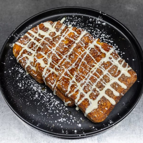 Three pieces of French toast drizzled with white icing and powdered sugar on a black plate.