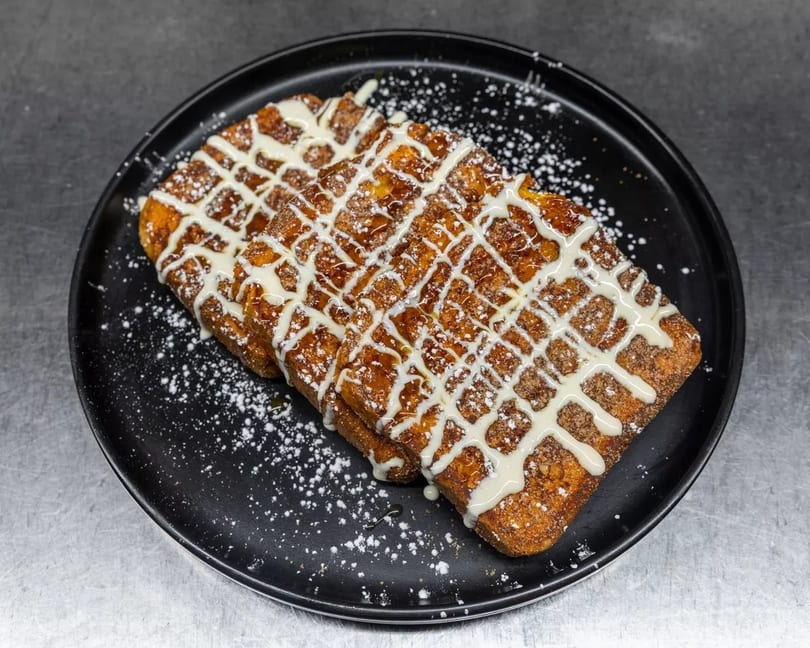 Three pieces of French toast drizzled with white icing and powdered sugar on a black plate.