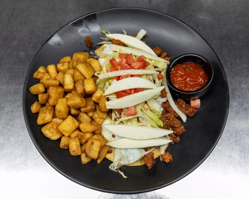 Breakfast plate with crispy home fries, huevos rancheros topped with cheese, lettuce, and tomatoes, and a side of salsa.