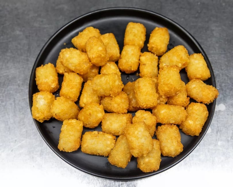 A black plate filled with golden-brown, crispy tater tots on a metallic surface.