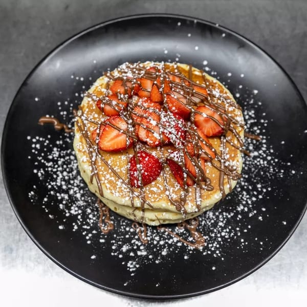 Stack of pancakes topped with strawberries, chocolate drizzle, and powdered sugar on a black plate.