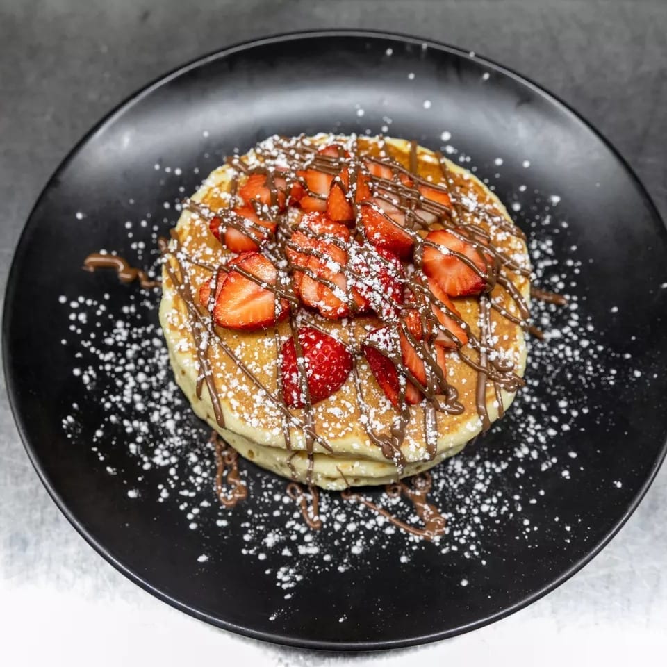 Stack of pancakes topped with strawberries, chocolate drizzle, and powdered sugar on a black plate.