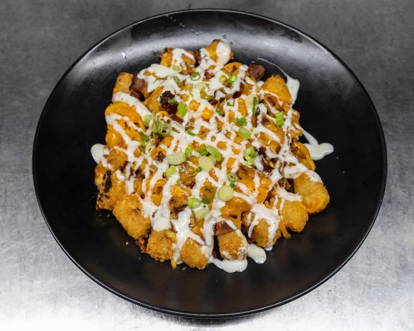 Loaded tater tots with melted cheese, bacon, ranch, and green onions on a black plate.