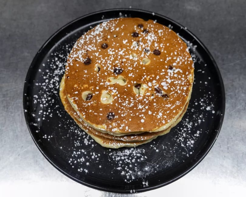 Stack of chocolate chip pancakes dusted with powdered sugar on a black plate.