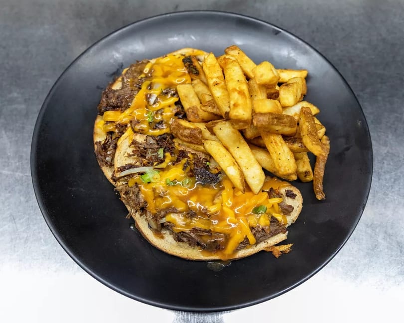 Open-faced cheesesteak sandwich with melted cheddar, green onions, and a side of golden fries.