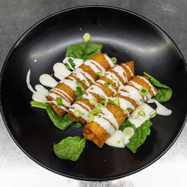 Crispy golden egg rolls drizzled with white sauce and garnished with green onions on a black plate.