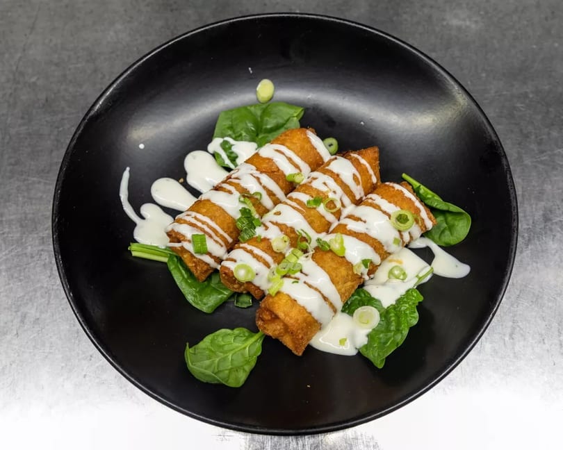 Crispy golden egg rolls drizzled with white sauce and garnished with green onions on a black plate.