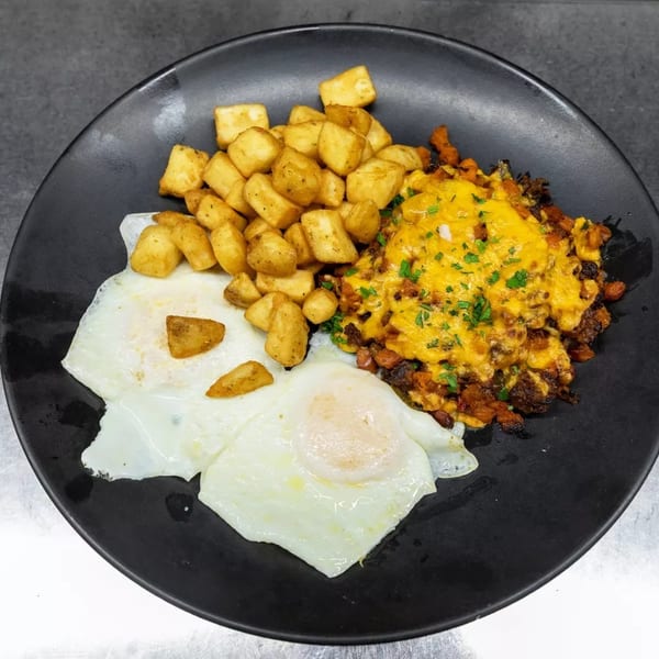 A black plate holds two fried eggs, crispy breakfast potatoes, and a cheesy chorizo hash.