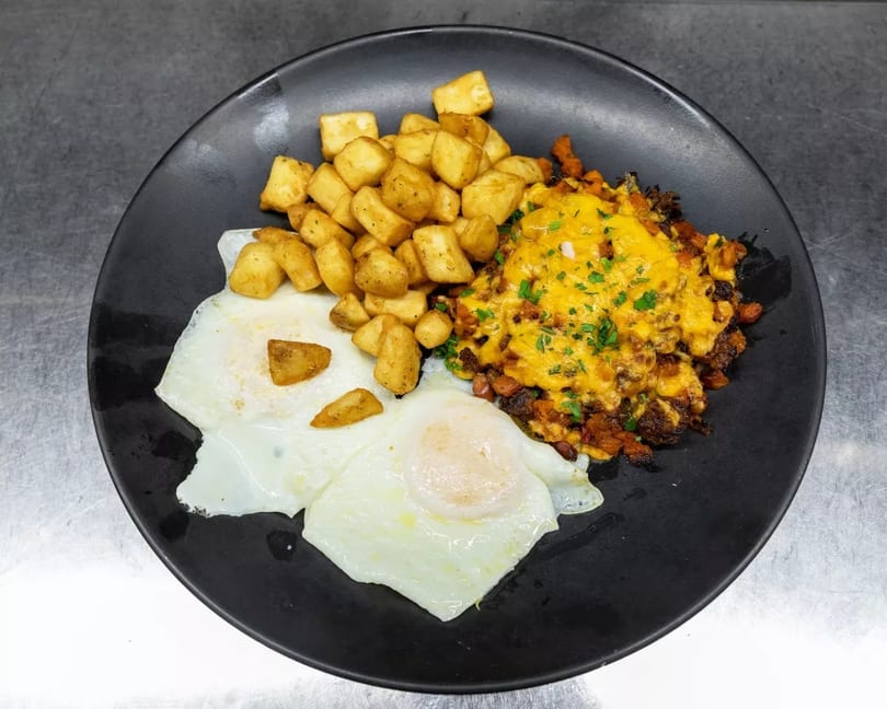 A black plate holds two fried eggs, crispy breakfast potatoes, and a cheesy chorizo hash.