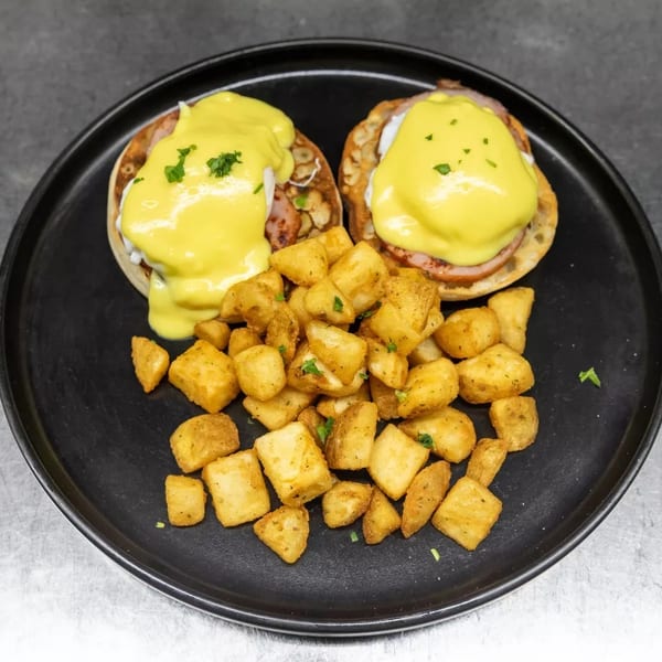 Two eggs Benedict with hollandaise sauce and a side of crispy home fries on a black plate.