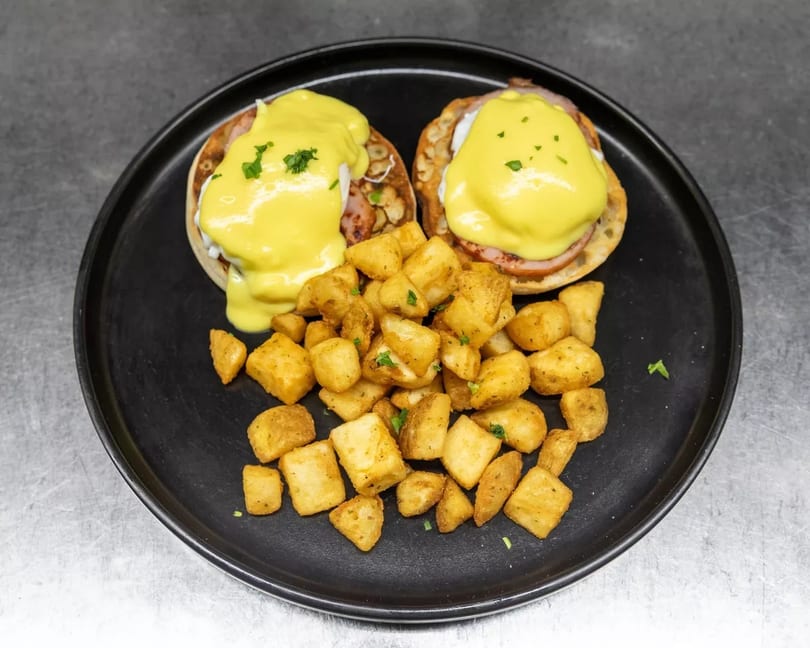 Two eggs Benedict with hollandaise sauce and a side of crispy home fries on a black plate.