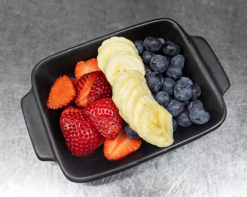 A black rectangular dish filled with fresh sliced strawberries, banana, and blueberries on a gray surface.